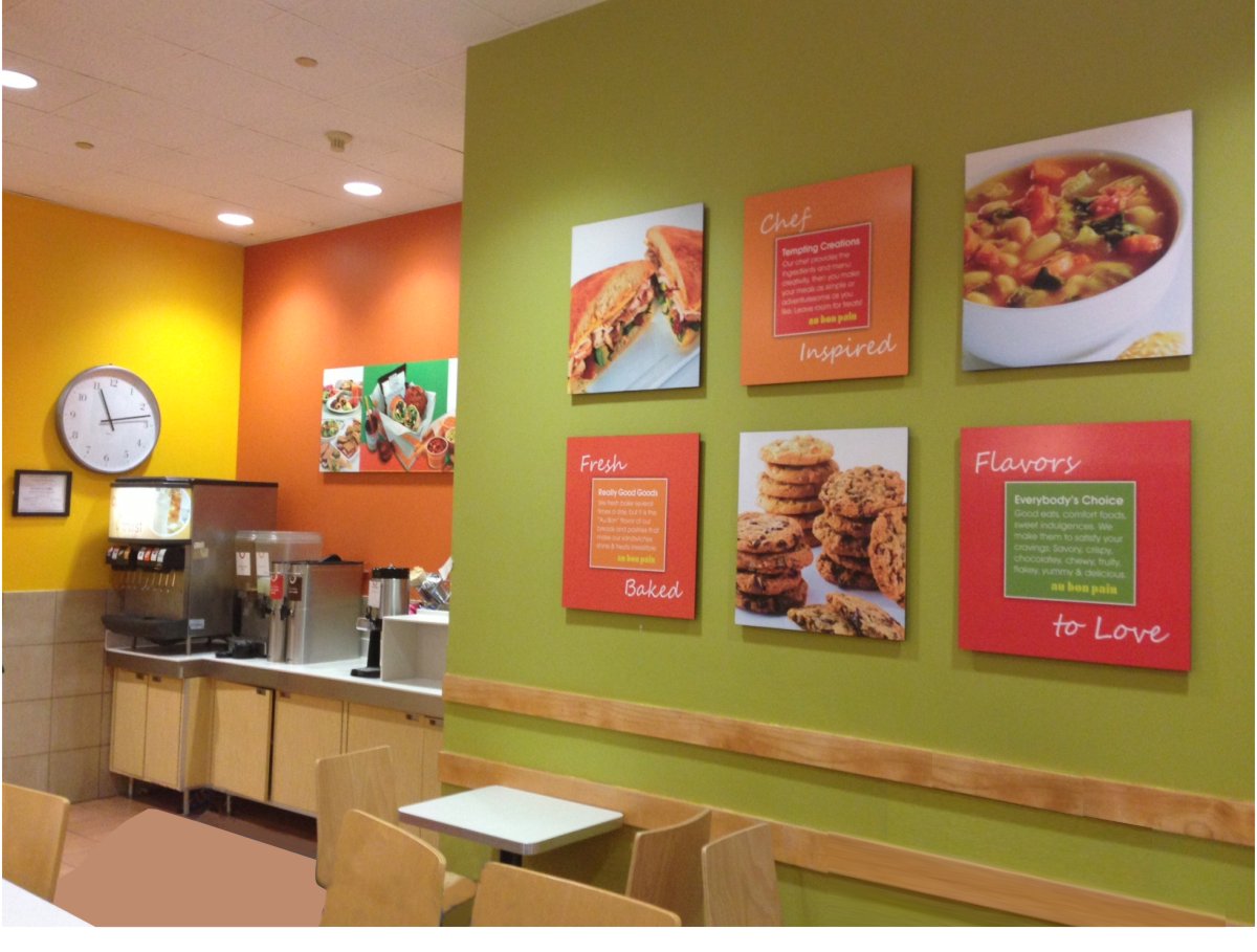 Color photo showing enhanced interior prototypical modular marketing graphic design system and painted wall finish color blocking scheme. From the left a yellow wall with a black and white clock centered; an orange wall with a horizontal catering poster and on the right a bright yellow green wall with a grid of six graphic poster elements, two high by three wide, twenty-four inch square photos of a sandwich top left cookies bottom center bowl of soup top right. In between there are orange brand promise text squares. Bottom left top center and bottom right saying fresh baked, taste tempting and flavors to love respectively.