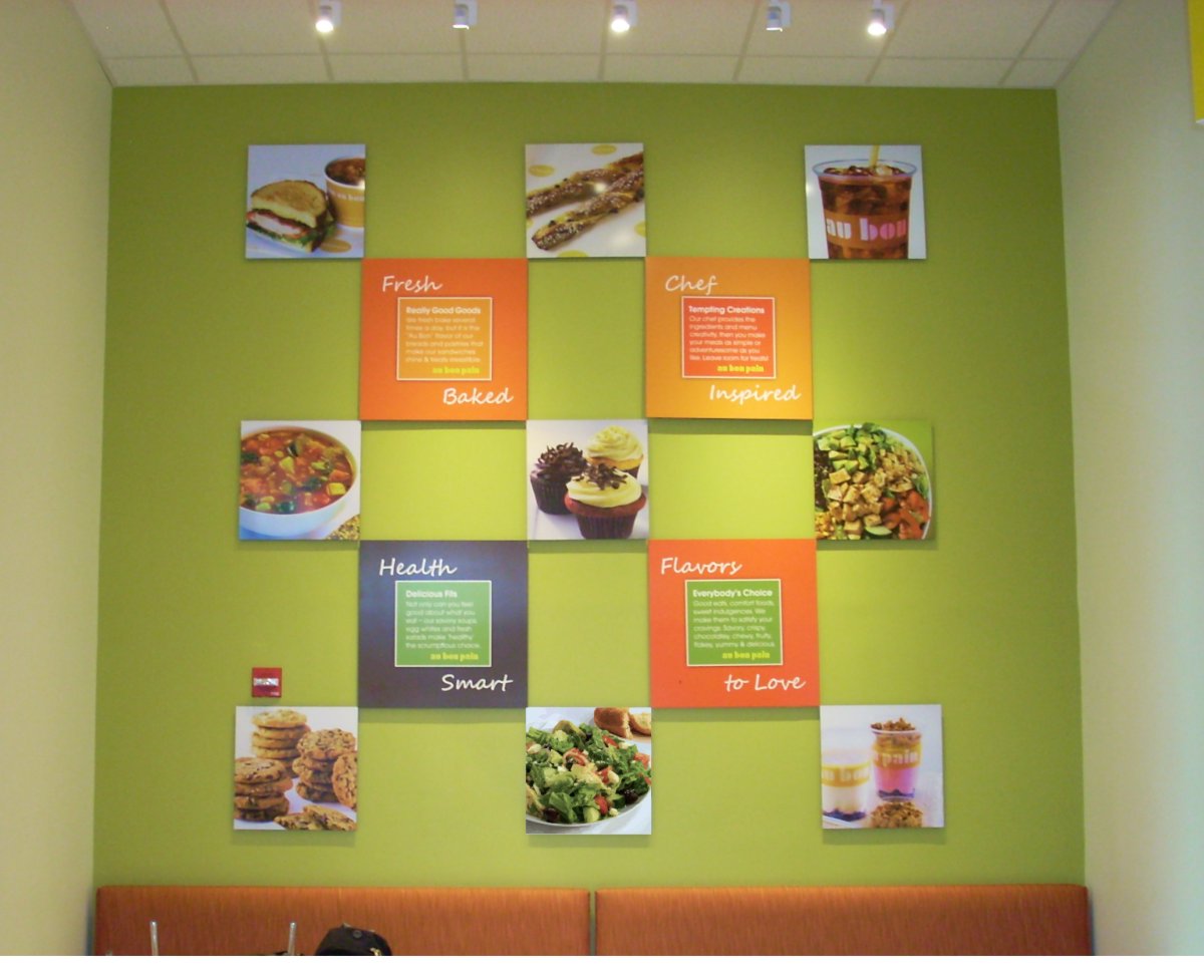 There is a grid of nine photos, three high and three wide featuring soup, sandwiches, salads, dessert cupcakes and cookies and breakfast yogurts. In between the photos are four orange green and brown brand promise text squares describing fresh baked, chef inspired, health smart and flavors to love attributes. These are mounted on a painted yellow green wall over orange fabric upholstered banquette seating. 